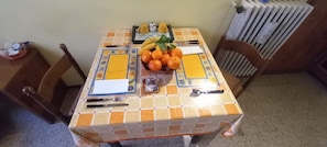 Une table à manger recouverte d’une nappe à motifs, un panier de fruits contenant des oranges et des bananes, et un petit récipient à sel.