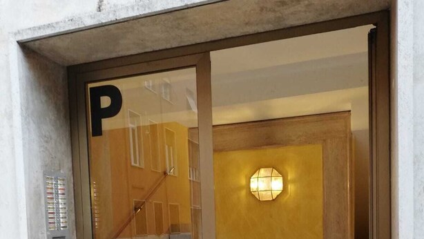 Ein Gebäudeeingang mit Glastür, gekennzeichnet mit einem 'P' für Parkplätze, einer Innentreppe und einer wandmontierten Leuchte.