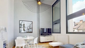 Un salon moderne doté d’une grande fenêtre, d’un coin repas avec des chaises blanches, d’un téléviseur à écran plat et d’un canapé gris.