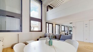 Un salon moderne avec une table ronde blanche, des chaises blanches, un canapé gris et une grande fenêtre avec des stores.