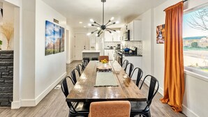 A dining area with a long wooden table, black chairs, a central light fixture, and a view of the kitchen and outdoors. - Family Retreat - Backyard Games + Movie Room - Sleeps 16 (Orem)