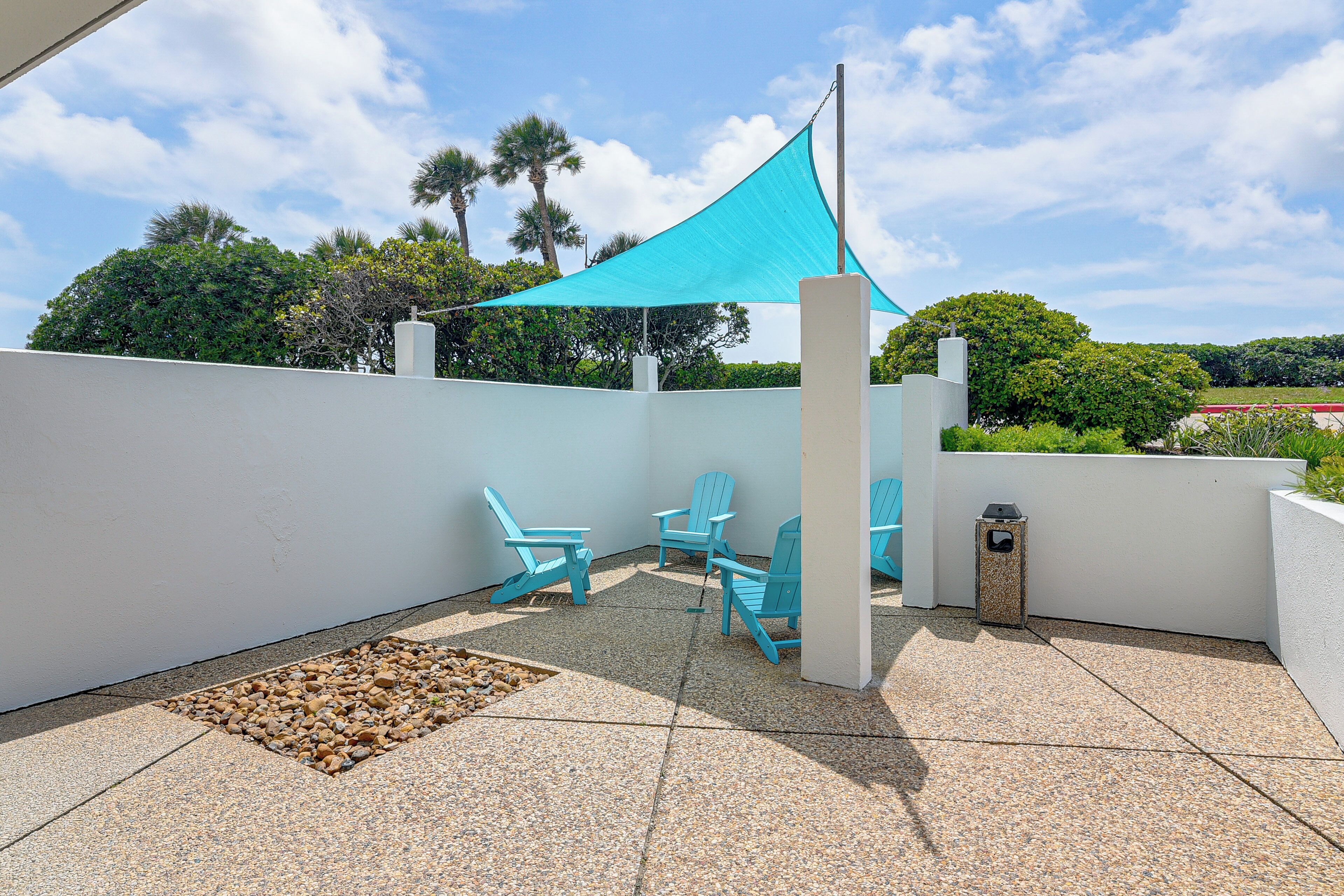 A patio with a blue shade sail, three blue Adirondack chairs, and a small trash bin.