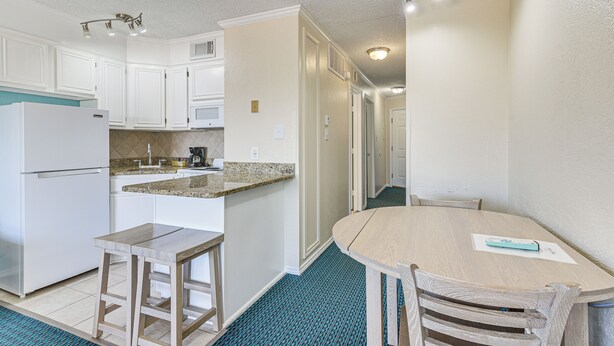 A compact kitchen with white cabinets, a built-in refrigerator, a granite countertop, and a wooden dining table with chairs. - Steps to Babes Beach: Condo w/ Pool in Galveston (Galveston)