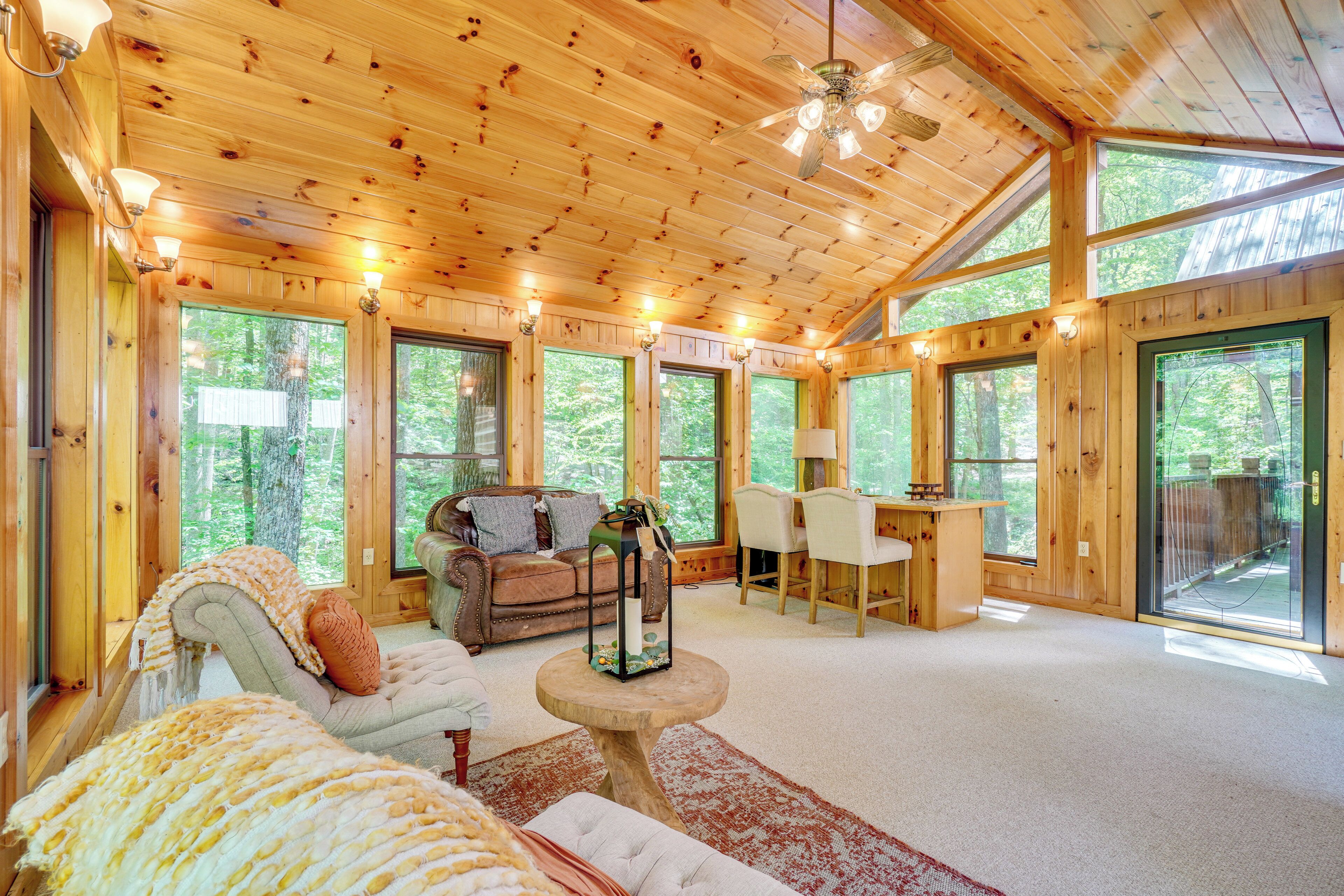 A cozy cabin interior with a wooden ceiling, large windows, a sofa, a dining table, and a fireplace.