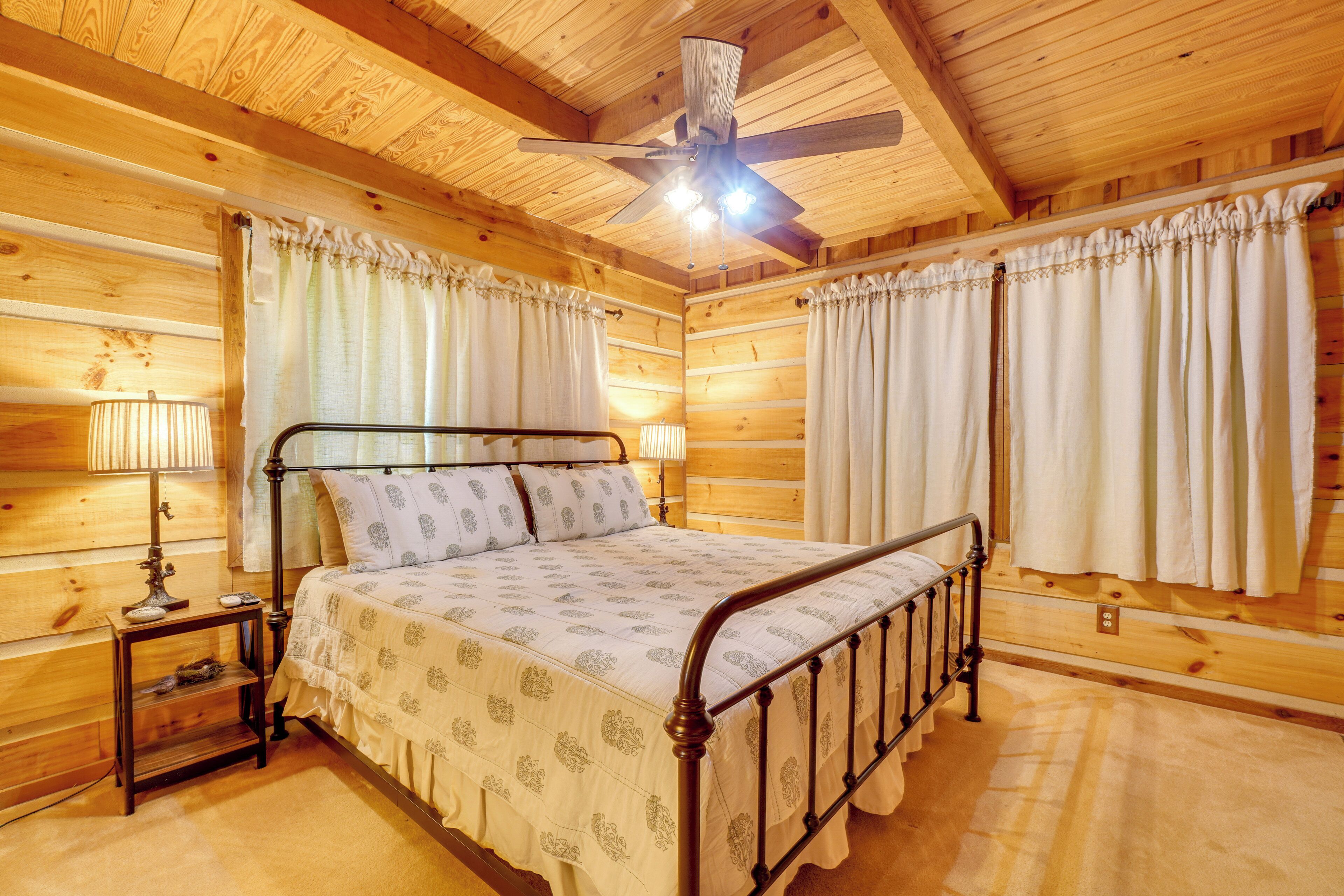 Dormitorio de cabaña de madera con cama, dos mesitas de noche, un ventilador de techo y cortinas.