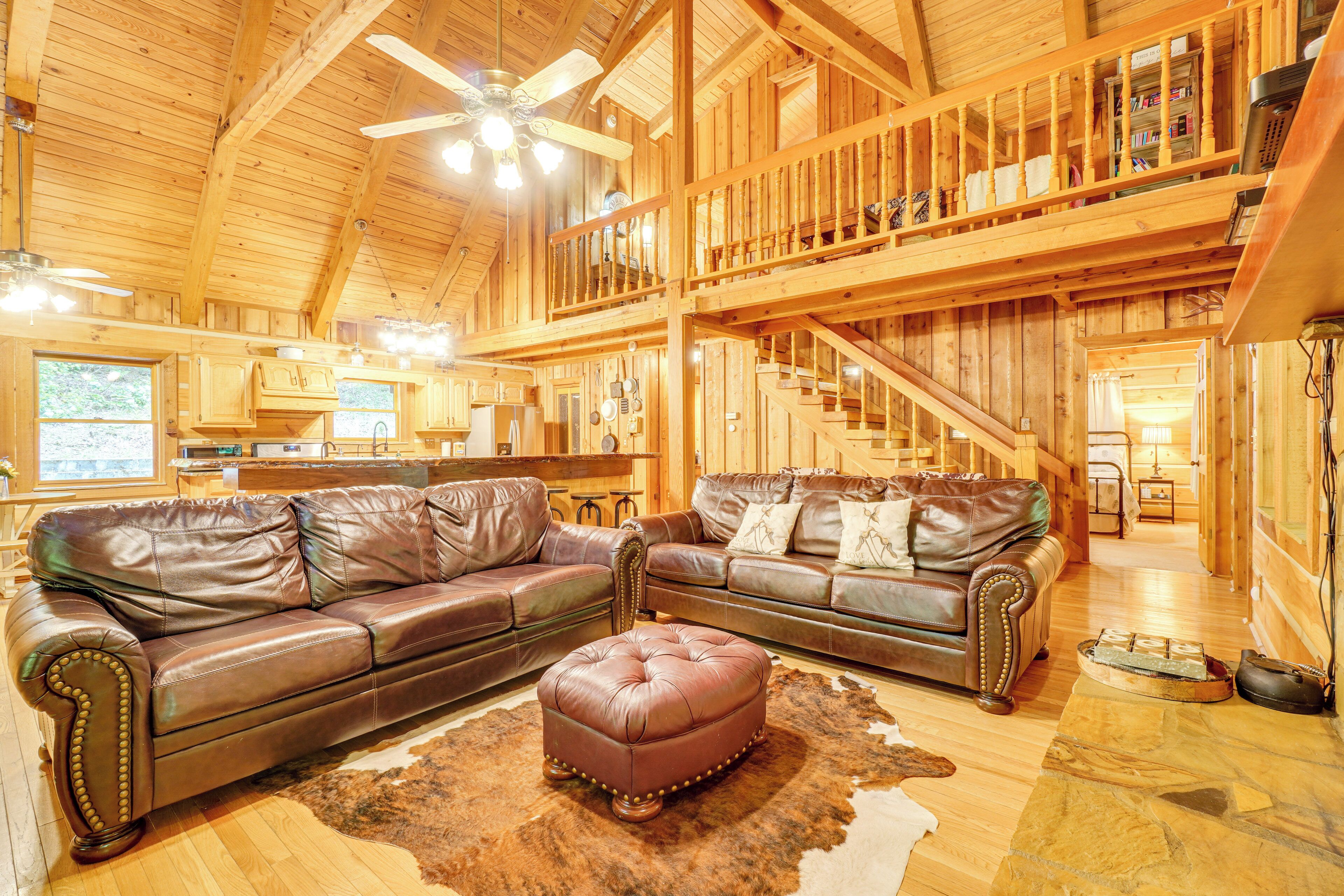 A spacious log cabin interior with a large living area, leather sofas, a central ottoman, and a staircase leading to an upper level.
