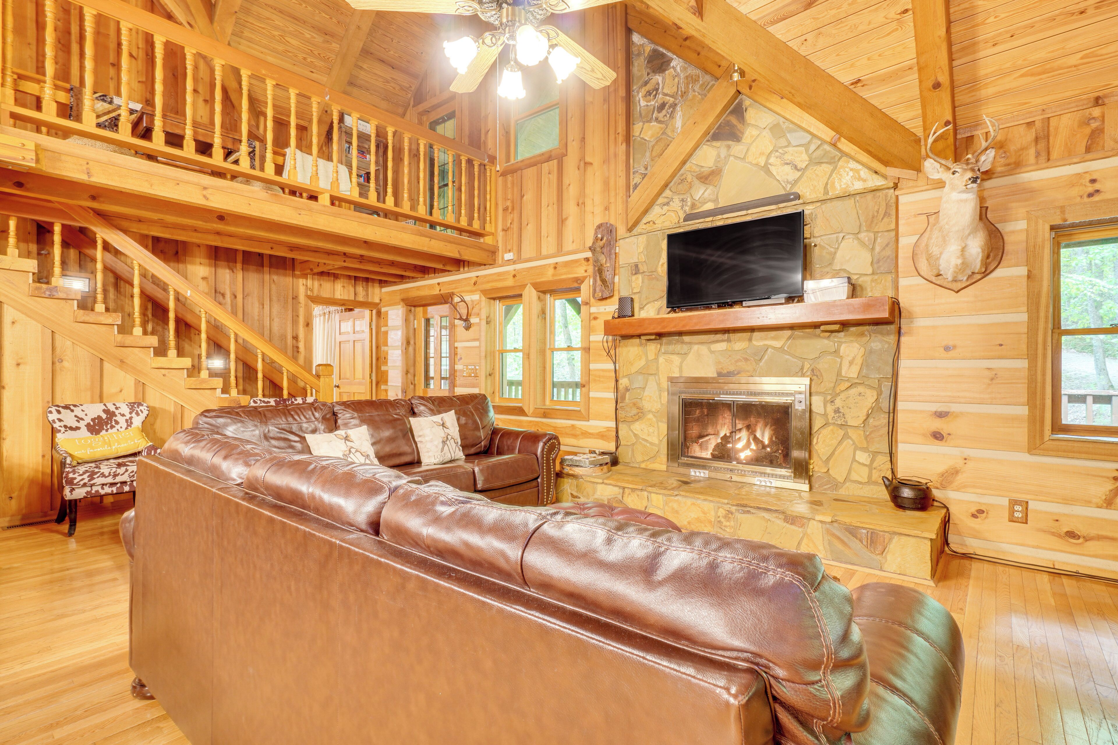 A log cabin interior with a stone fireplace, leather sofa, wooden staircase, and a mounted deer head.