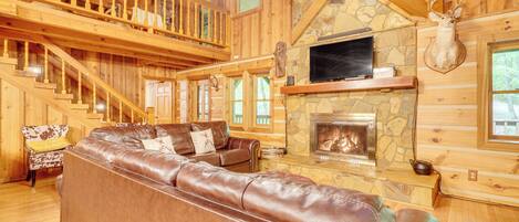 A log cabin interior with a stone fireplace, leather sofa, wooden staircase, and a mounted deer head.