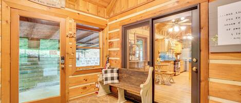 Interior de una cabaña de madera con piso de piedra, un banco y una puerta de vidrio que da a una zona de cocina.