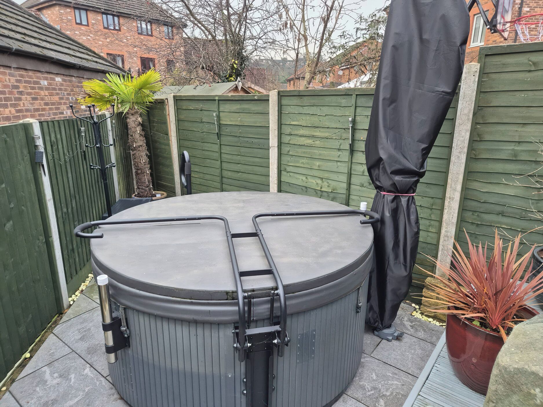 A hot tub with a ladder, surrounded by a green fence, a potted plant, and a brick house in the background.