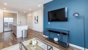 A modern kitchen with a built-in refrigerator, a dining area with a glass-top table, and a living room with a mounted TV and a blue accent wall. - Landing | Chic 1BD, Gym, Pool (Fate)