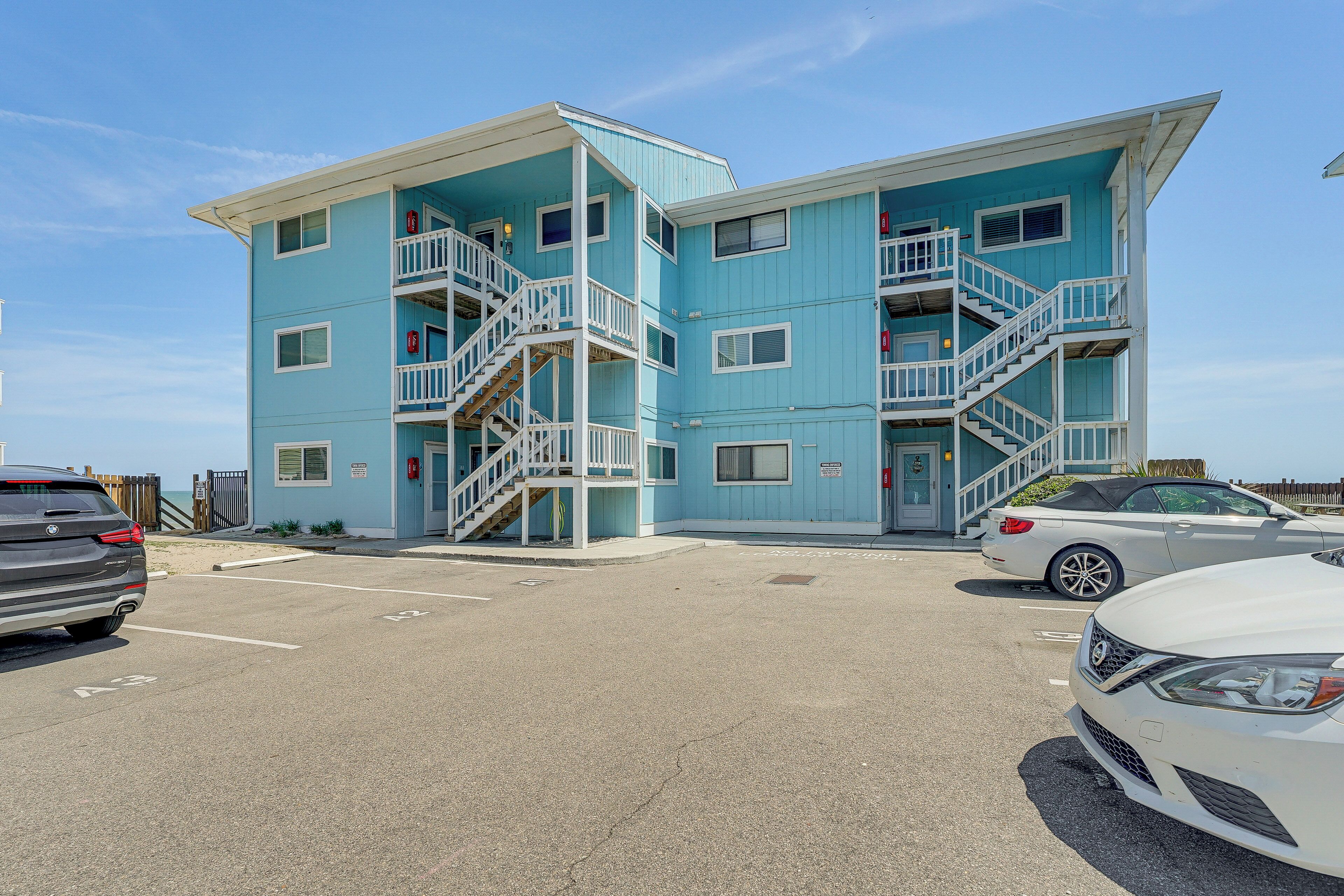 A multi-story building with a light blue exterior, white railings, and multiple balconies. There are cars parked in front, and a clear sky above.