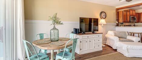A dining area with a wooden table, four chairs, and a vase of flowers. There is a kitchen with white cabinets, a microwave, and a ceiling fan.
