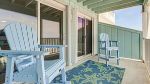 A covered outdoor area with two Adirondack chairs, a starfish-patterned rug, and a sliding glass door leading to a covered patio. - Sandy Oceanfront 2nd FL 1BR W/pool at The Riggings (Kure Beach)