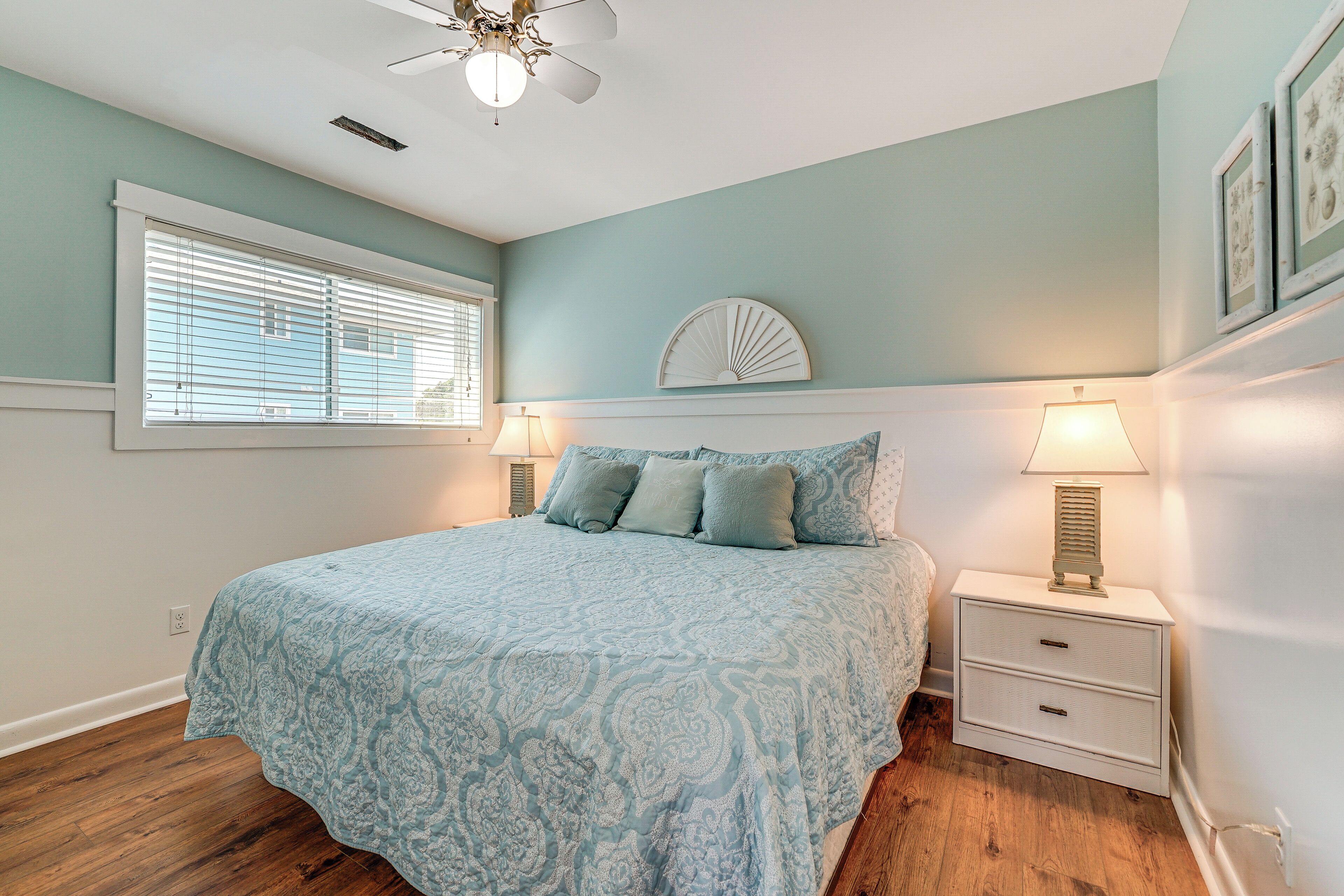 A bedroom with a bed, two bedside tables, a ceiling fan, a window with blinds, and a wall-mounted decorative piece.
