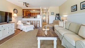 A living room with a TV, white furniture, a wooden coffee table, and a kitchen in the background. - Sandy Oceanfront 2nd FL 1BR W/pool at The Riggings (Kure Beach)