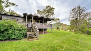 Una casa de dos pisos con balcón, rodeada de exuberante vegetación y colinas al fondo.