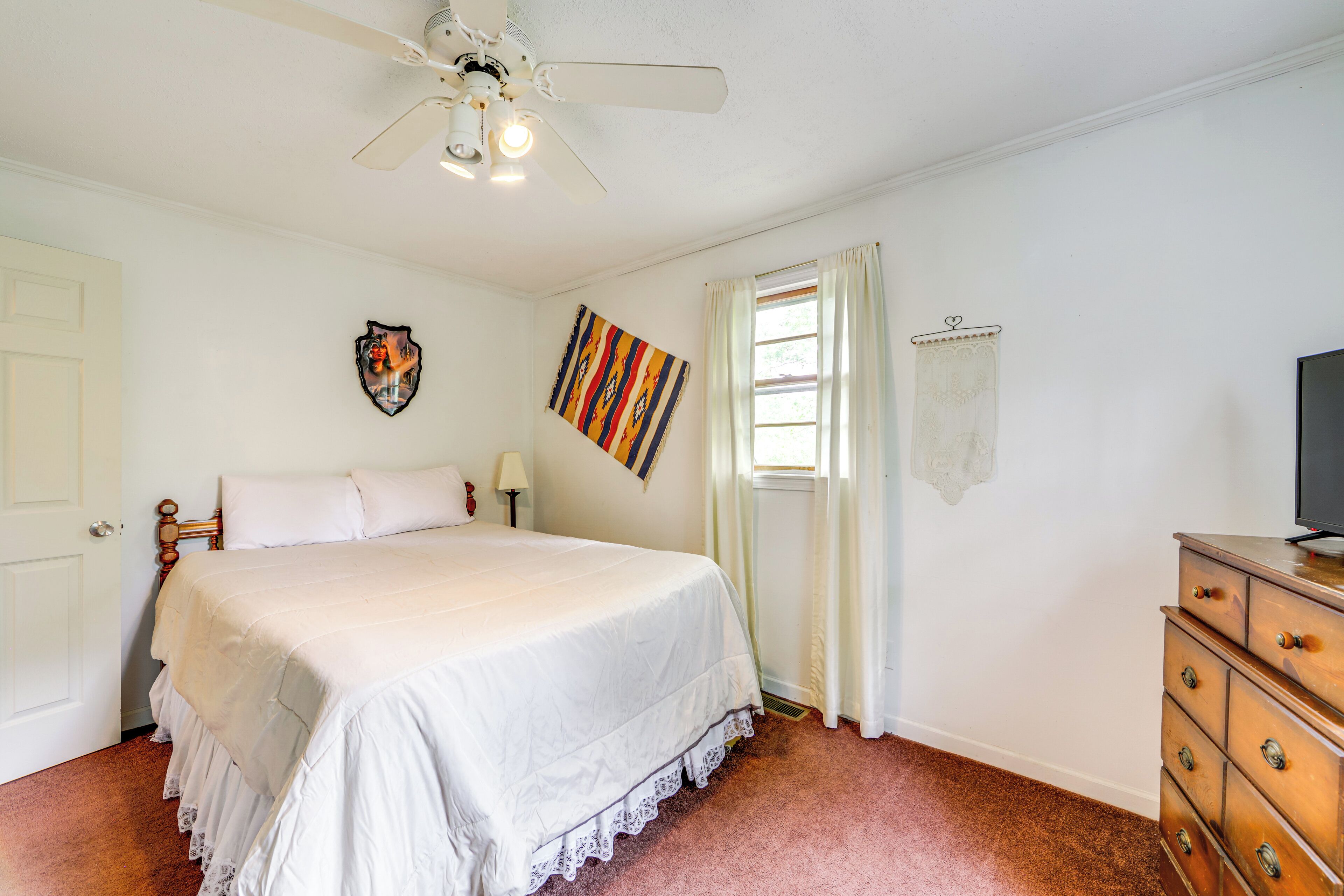 Un dormitorio con cama, cómoda, ventilador de techo, ventana con cortinas y una obra de arte colgada en la pared.