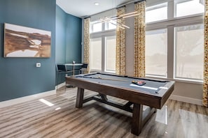 Une table de billard dans une pièce avec un mur bleu, un tableau et une fenêtre avec des rideaux.