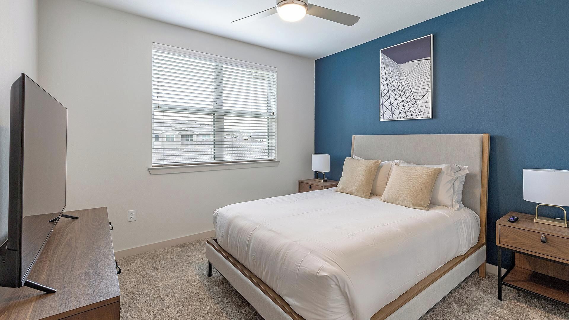 A bedroom with a bed, two bedside tables, a nightstand, a ceiling fan, a window with blinds, and a wall-mounted artwork.