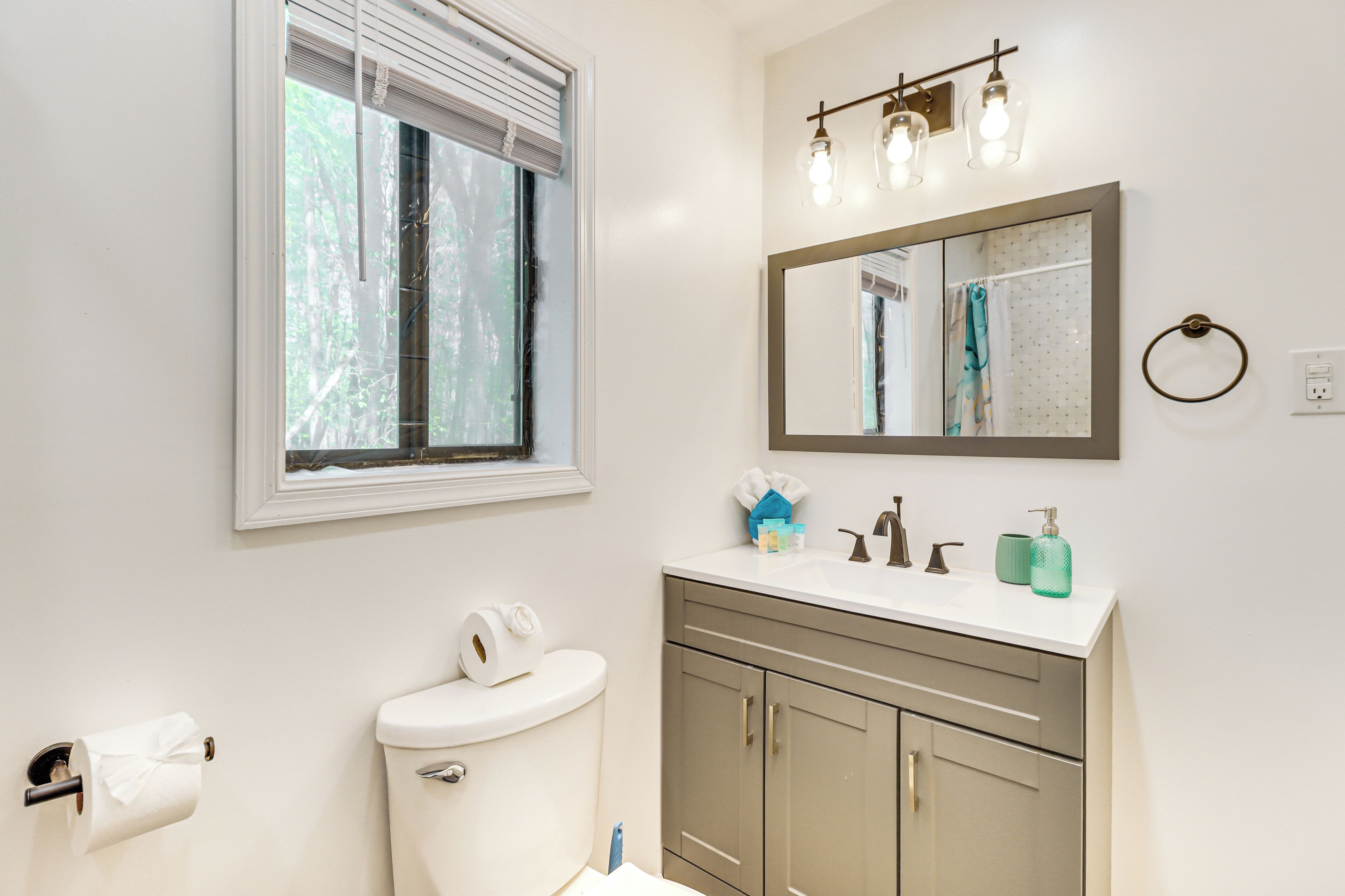 A bathroom with a toilet, sink, and mirror. There is a window with blinds, a towel rack, and a light fixture above the mirror.