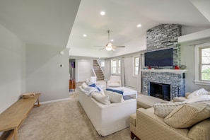 A spacious living room with a stone fireplace, a ceiling fan, and a large TV mounted above it.