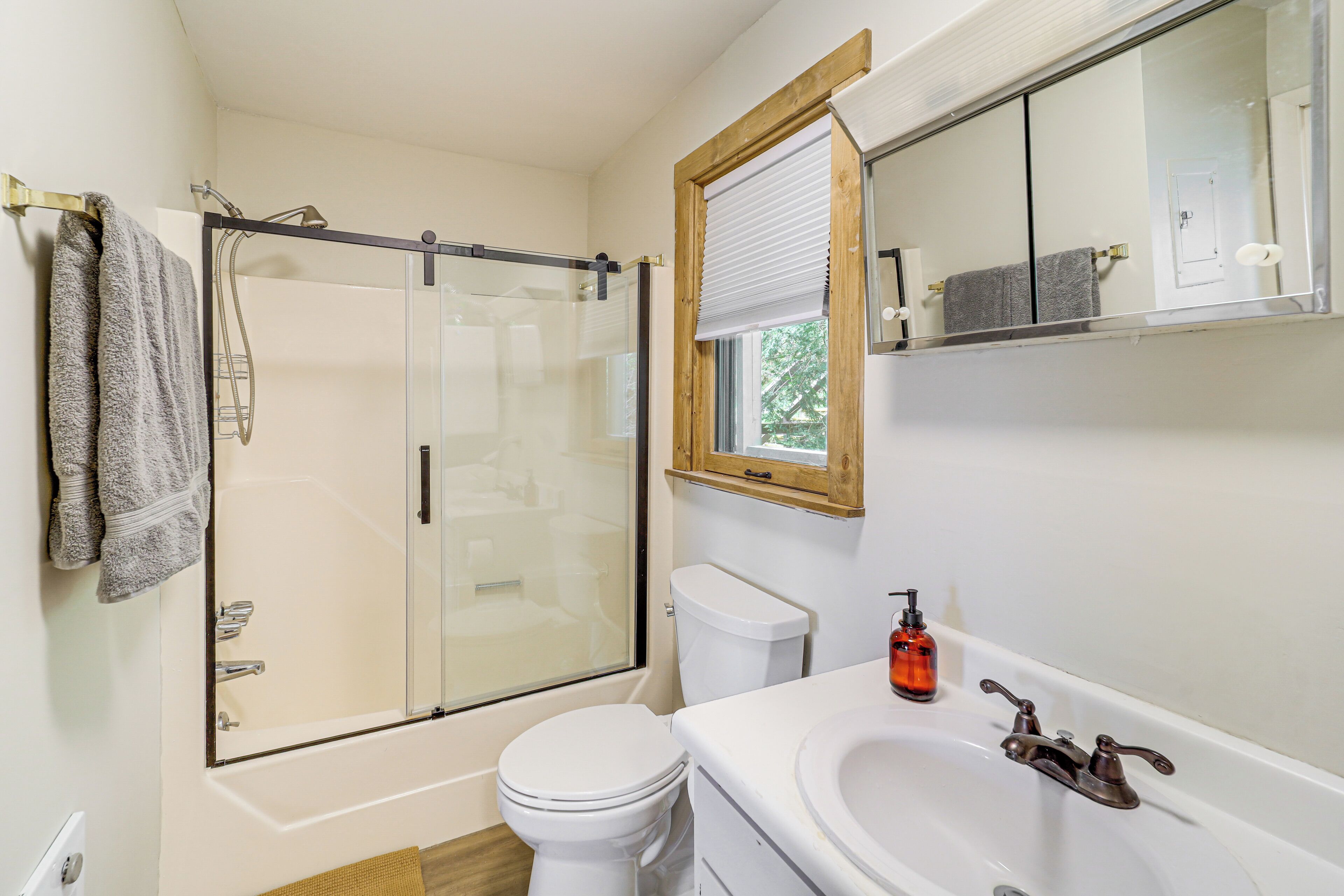 A bathroom with a glass-enclosed shower, a white toilet, a sink with a mirror, and a window with a wooden frame.