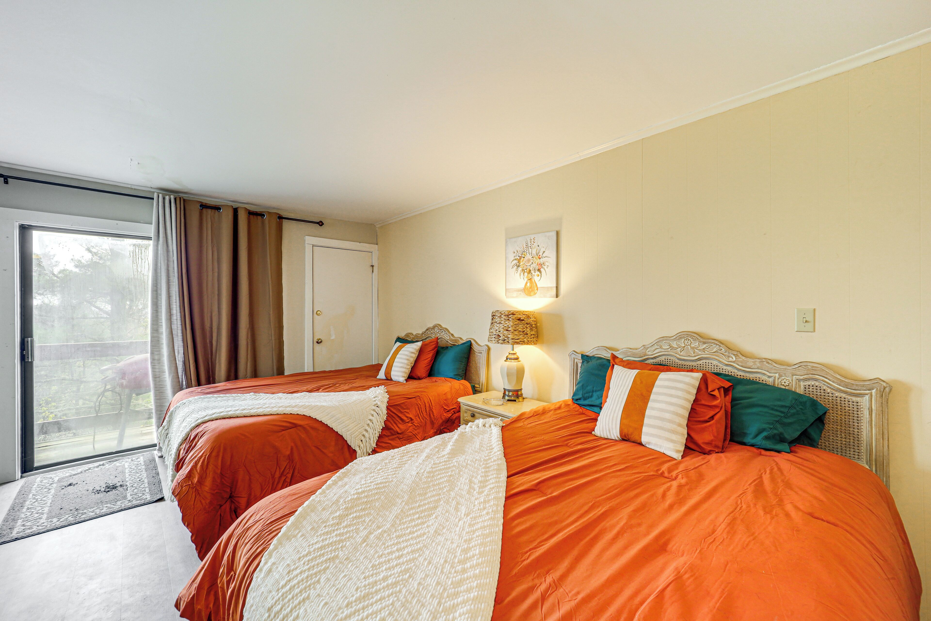 A hotel room with two beds, a window with curtains, a nightstand with a lamp, and a wall-mounted artwork.