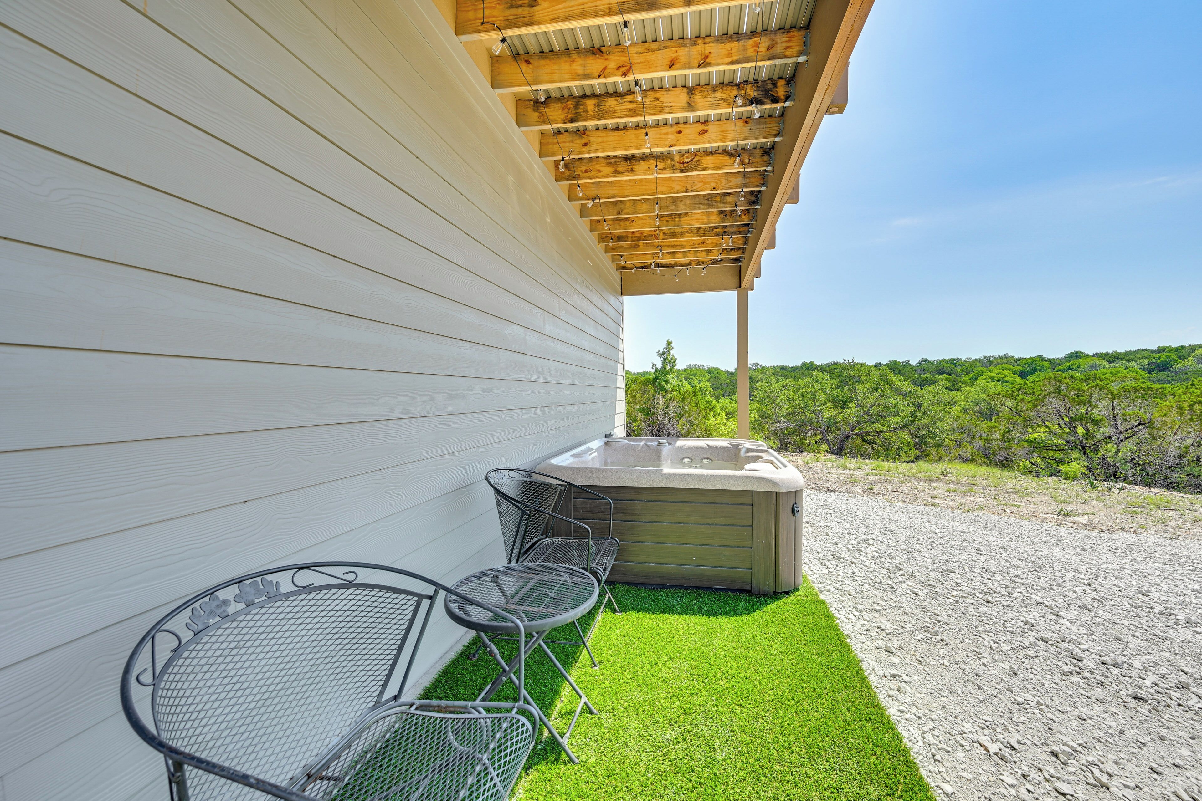 A covered outdoor area with a hot tub, two metal chairs, and a view of a green landscape.