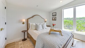 A bedroom with a large bed, a bedside table with a lamp, a window with a view of greenery, and a door with a brass handle. - Hot Tub & Valley Views: Hidden Gem in Texas (Valley Mills)