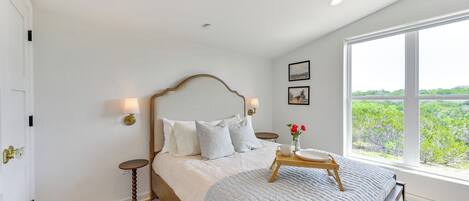 A bedroom with a large bed, a bedside table with a lamp, a window with a view of greenery, and a door with a brass handle.