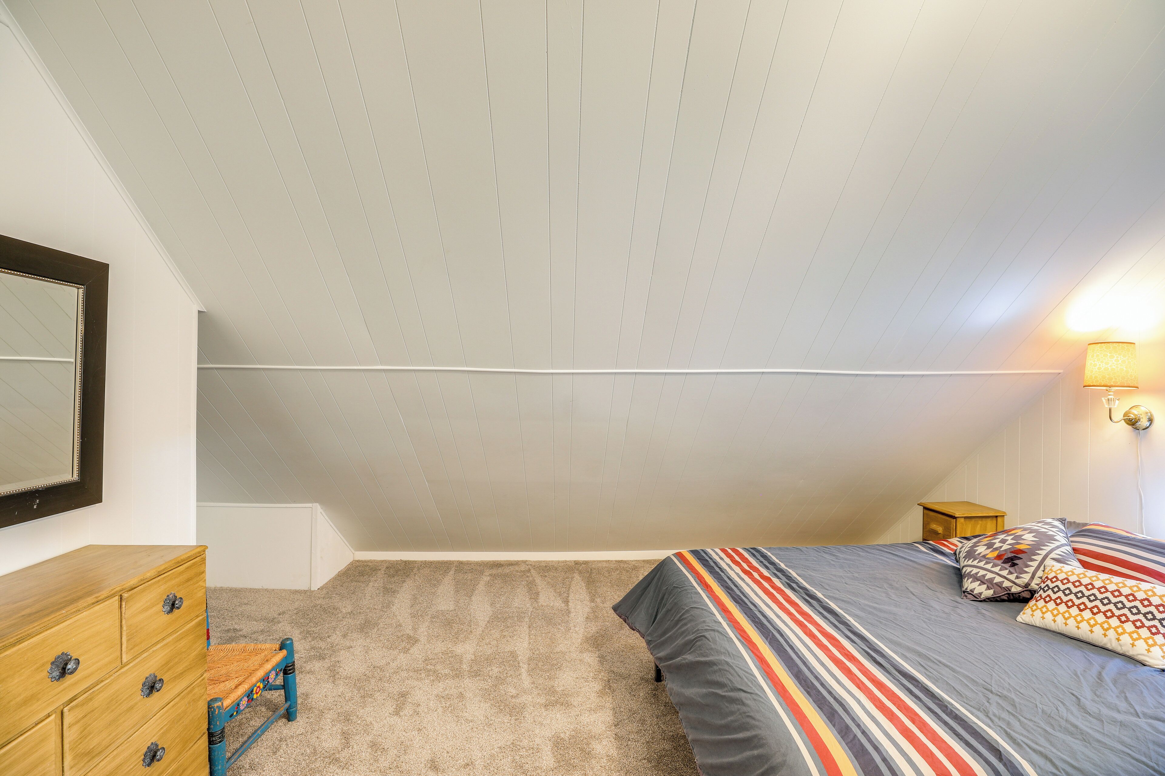 Un dormitorio con techo inclinado, cómoda de madera, una silla pequeña, cama con ropa de cama a rayas y mesita de noche con lámpara.