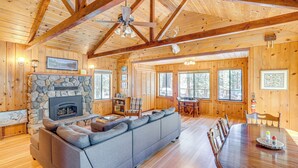 A cozy cabin interior with a stone fireplace, a gray sofa, wooden dining table, and large windows.