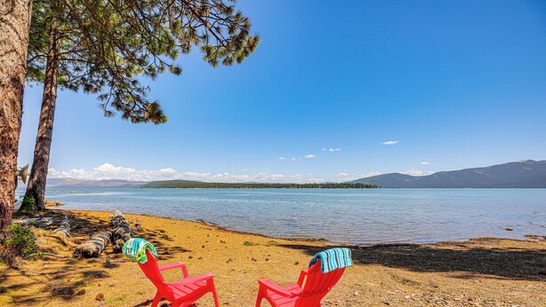 Two red chairs with blankets on a sandy beach by a lake, with a tree and mountains in the background.
