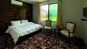 A hotel room with a bed, two chairs, a small table, a window with curtains, and a TV on the wall.