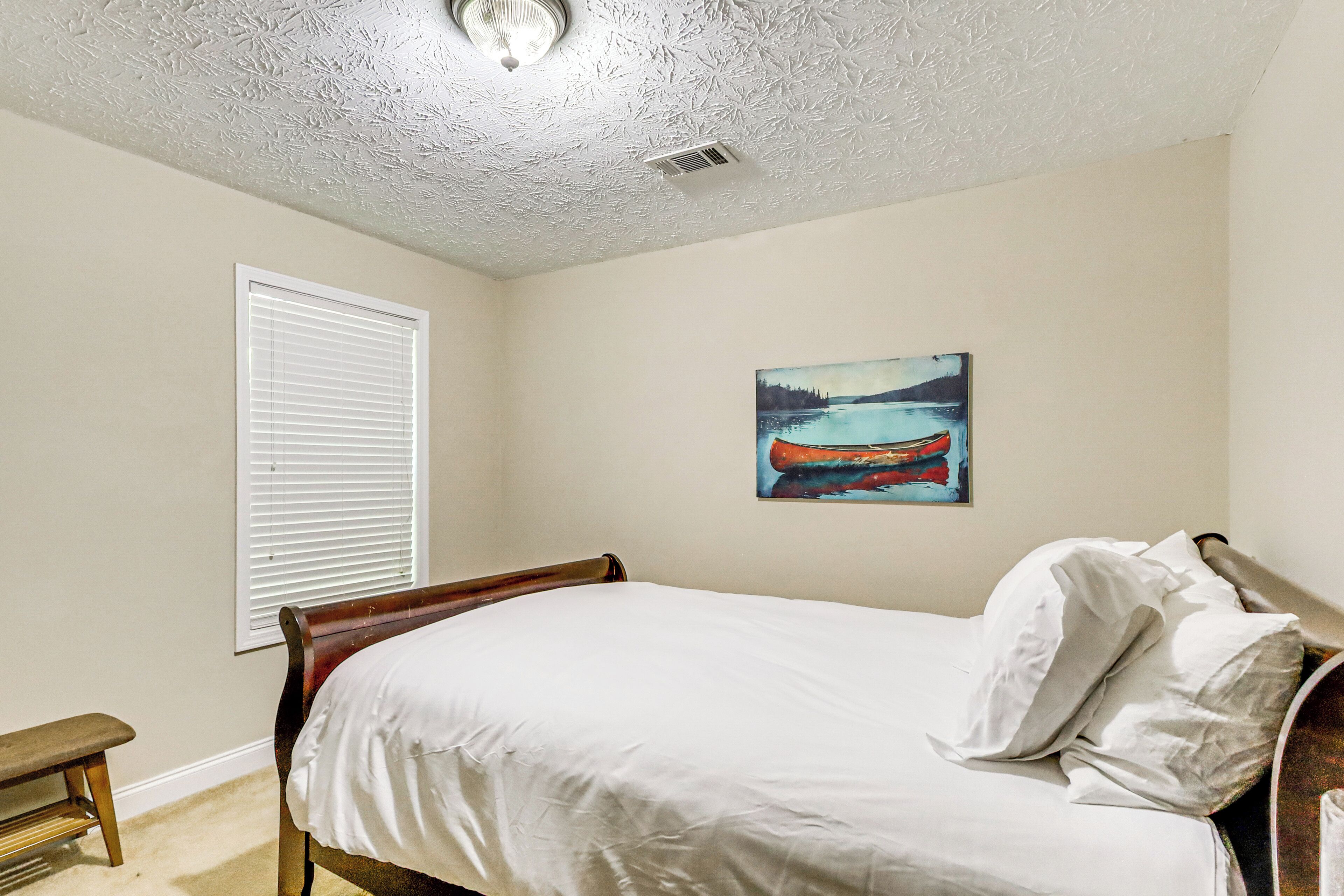 A bedroom with a bed, a bench, a window with blinds, and a painting on the wall.