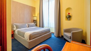 Quarto de hotel com cama, cadeira, escrivaninha, abajur e janela com cortinas.
