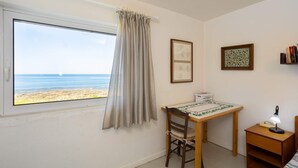 A room with a window overlooking a beach, a wooden desk with a tablecloth, a chair, a small bookshelf, and a lamp. - Agrilos Seafront Serenity - Waterfront Oasis (Trifylia)