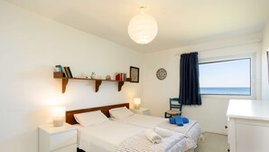 A hotel room with two beds, a nightstand with a lamp, a shelf with books and decorative items, a window with a view, and a chair. - Agrilos Seafront Serenity - Waterfront Oasis (Trifylia)