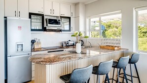 A modern kitchen with white cabinets, a built-in microwave, a refrigerator, a marble countertop, and a large window offering a view of greenery. - Ocean View 2 bed Apt in Camps Bay Skyline Heights (Cape Town)
