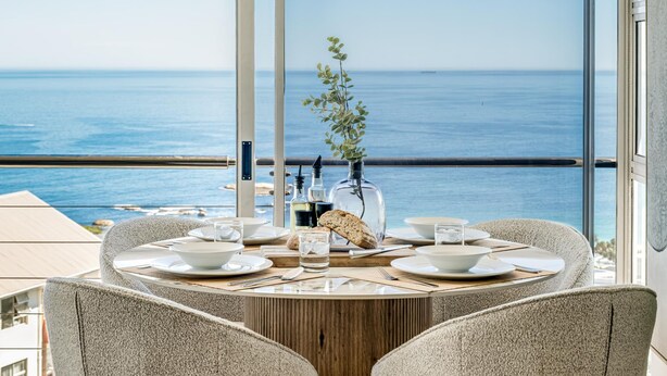 A dining table with chairs, a vase with a plant, and a bottle of water. - Ocean View 2 bed Apt in Camps Bay Skyline Heights (Cape Town)
