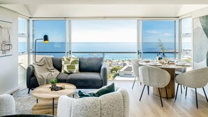 A modern living room with a large window overlooking the sea, a round wooden table, and a gray sofa. - Ocean View 2 bed Apt in Camps Bay Skyline Heights (Cape Town)