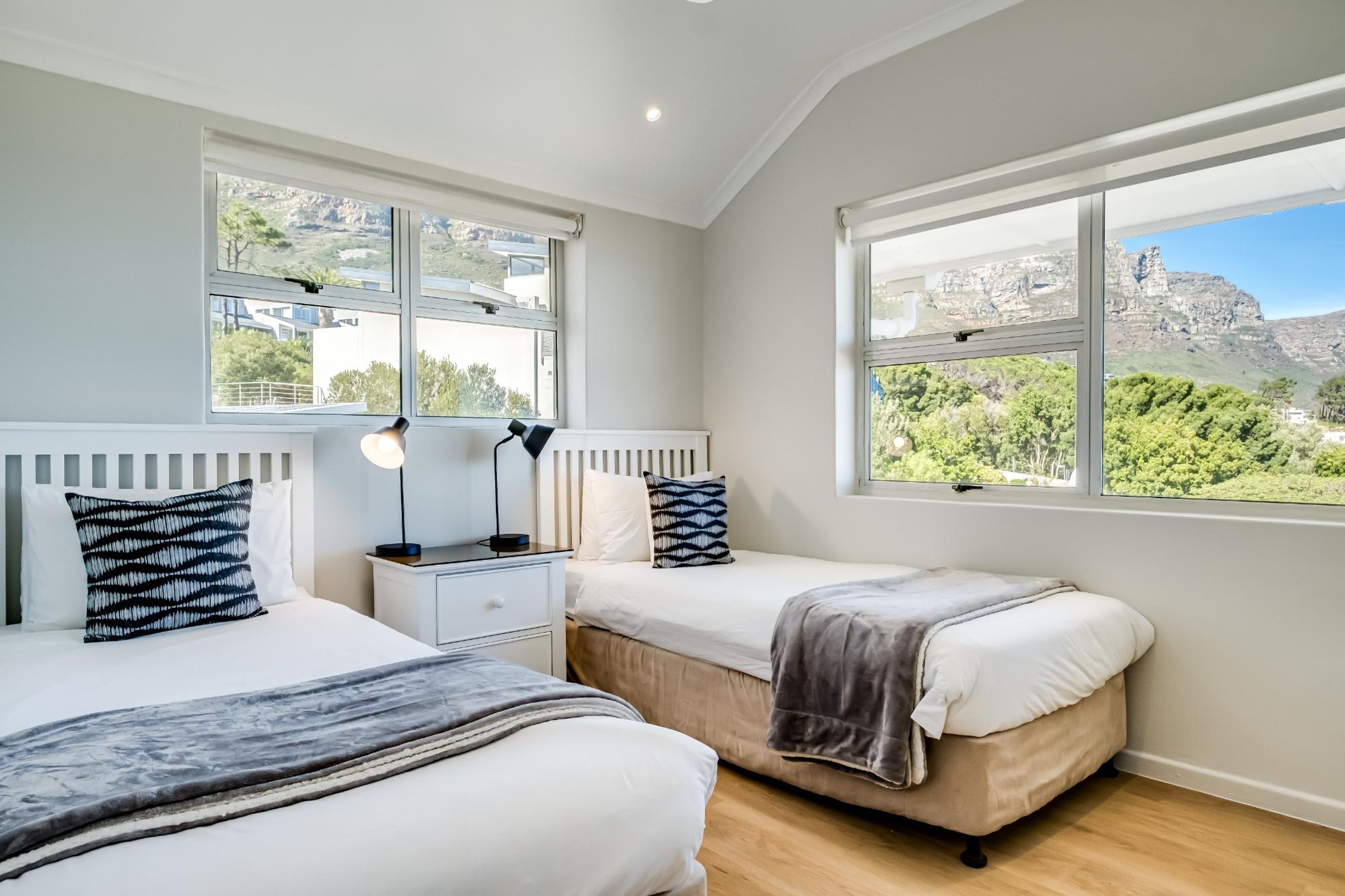 A bedroom with two beds, a nightstand, and a lamp. There are large windows with views of greenery and a mountain.