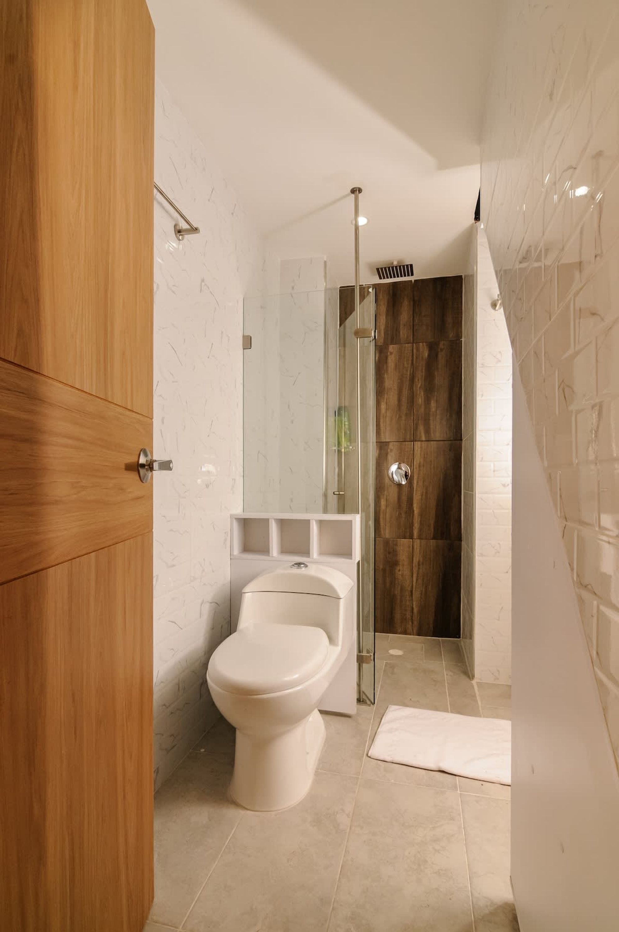 A modern bathroom with a wooden vanity, a white toilet, a glass shower enclosure, and a white tiled floor.