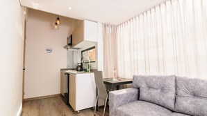 A compact living space with a kitchenette, a grey sofa, and a window with sheer curtains. - NOK Amazing Duplex in Parque 93 (Bogotá)