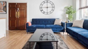A living room with a blue sofa set, a round wall clock, a small table, and a door with a brass handle. - Stunning Apartment in the ❤️ of Mayfair ❄️ Air Conditioning | Sleeps 5 (Greater London)