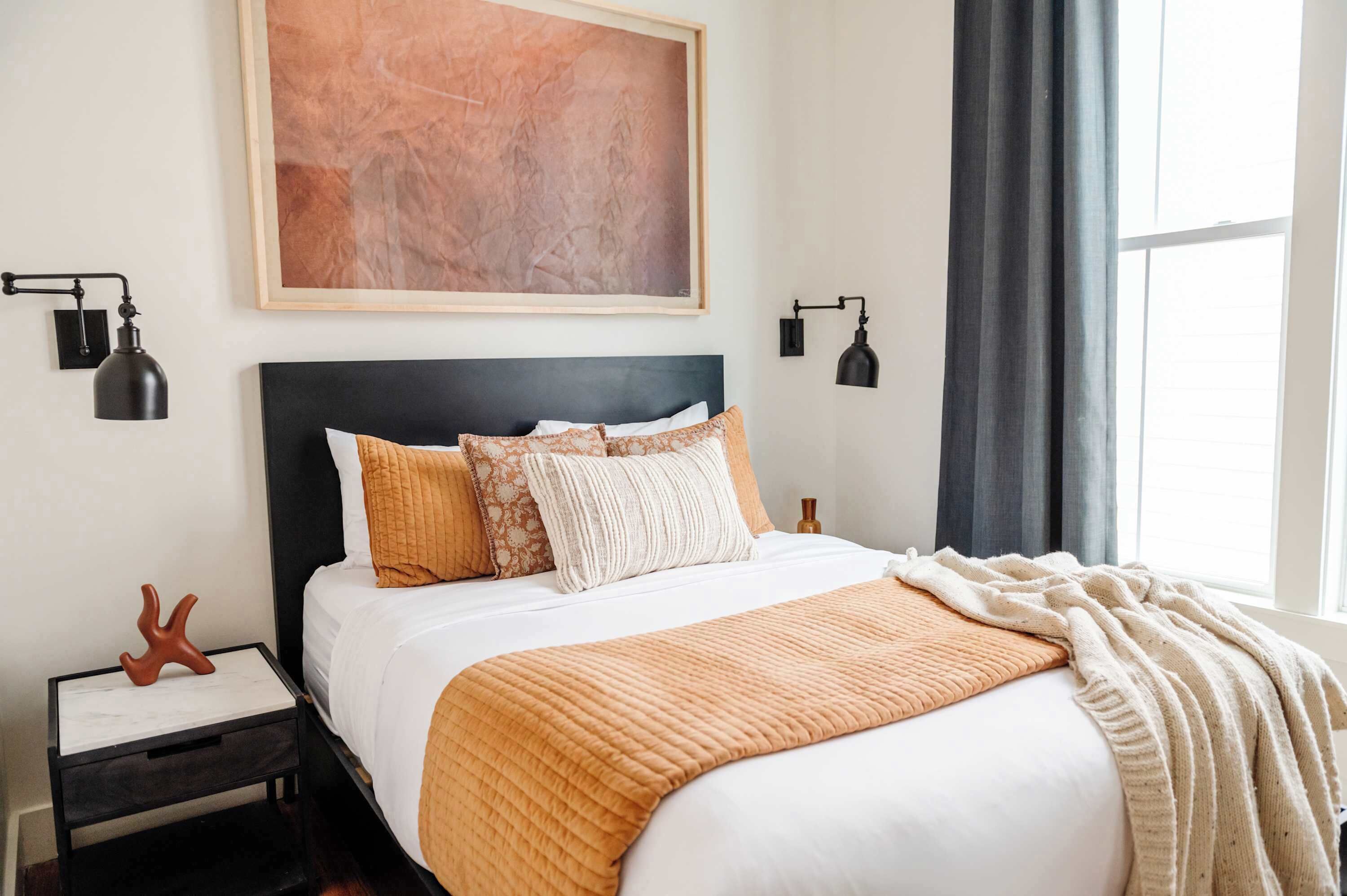 A neatly made bed with a white and orange quilt, a dark headboard, and a large abstract painting above it. There are two bedside lamps and a small decorative object on the nightstand.