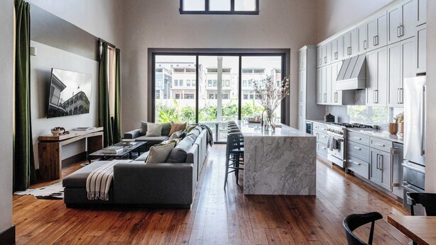 A modern kitchen with a large island, wooden flooring, and a view of buildings through large windows. - The 1300 | 2 Designer Homes, Walk to St Charles (New Orleans)