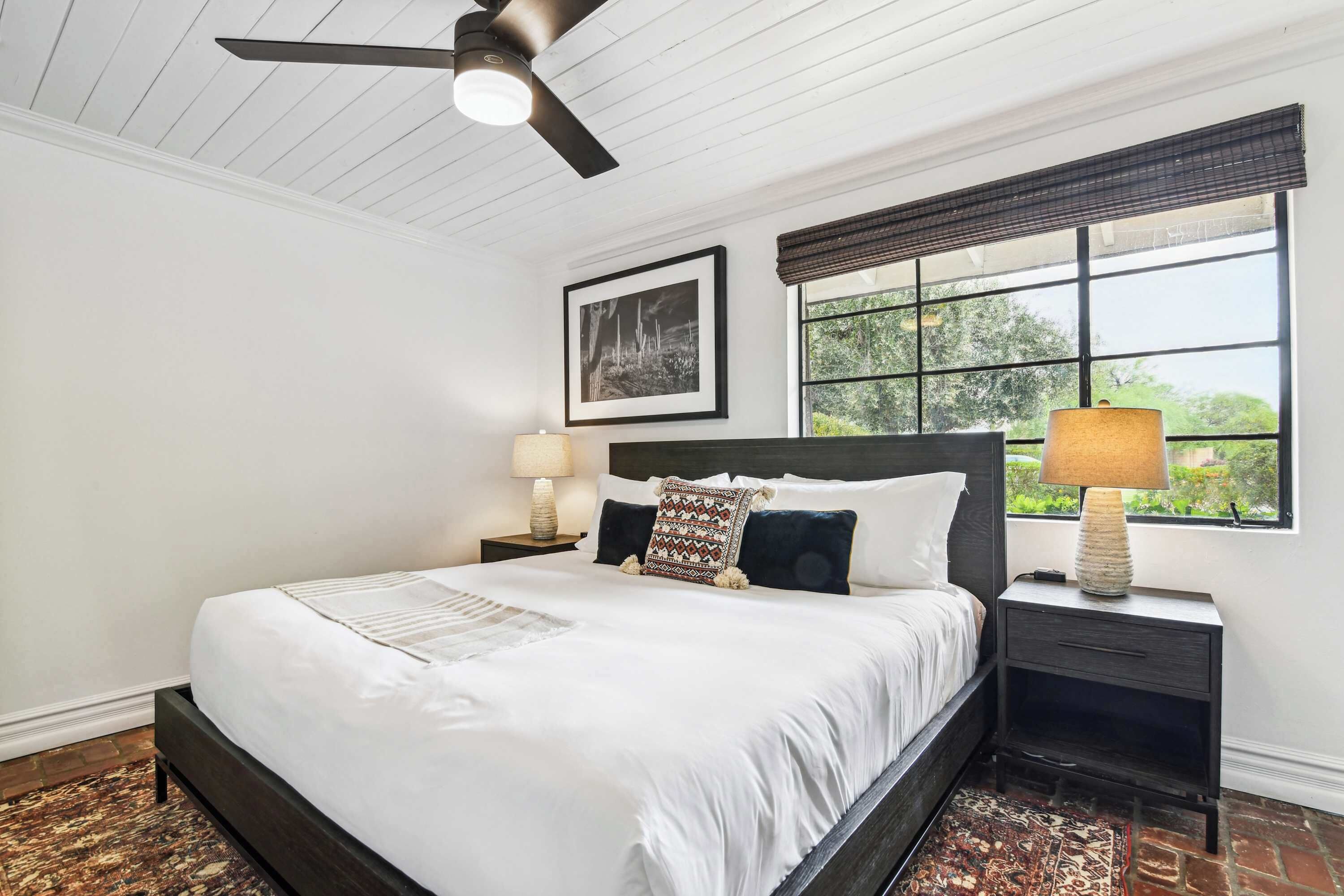 A bedroom with a large bed, a ceiling fan, a window with a view, a nightstand with a lamp, and a framed picture on the wall.