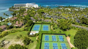 Oversigtsbillede af en golfbane med tennisbaner, omgivet af frodig vegetation og et hotel i baggrunden.